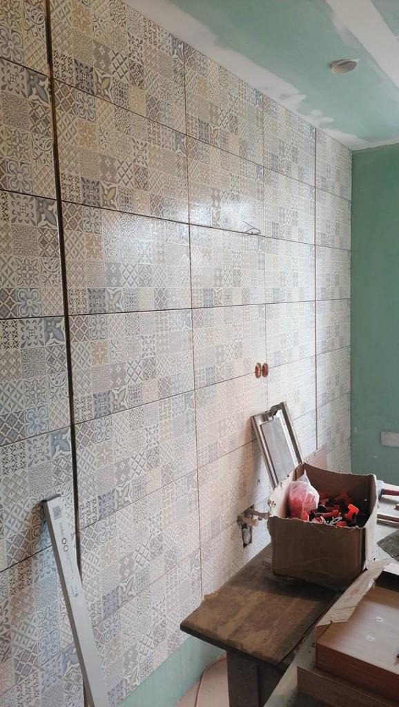 Ściana łazienki wyłożona patchworkowymi płytkami ceramicznymi w odcieniach szarości i beżu, widoczne narzędzia glazurnicze i karton z klinami do płytek na drewnianym stole.