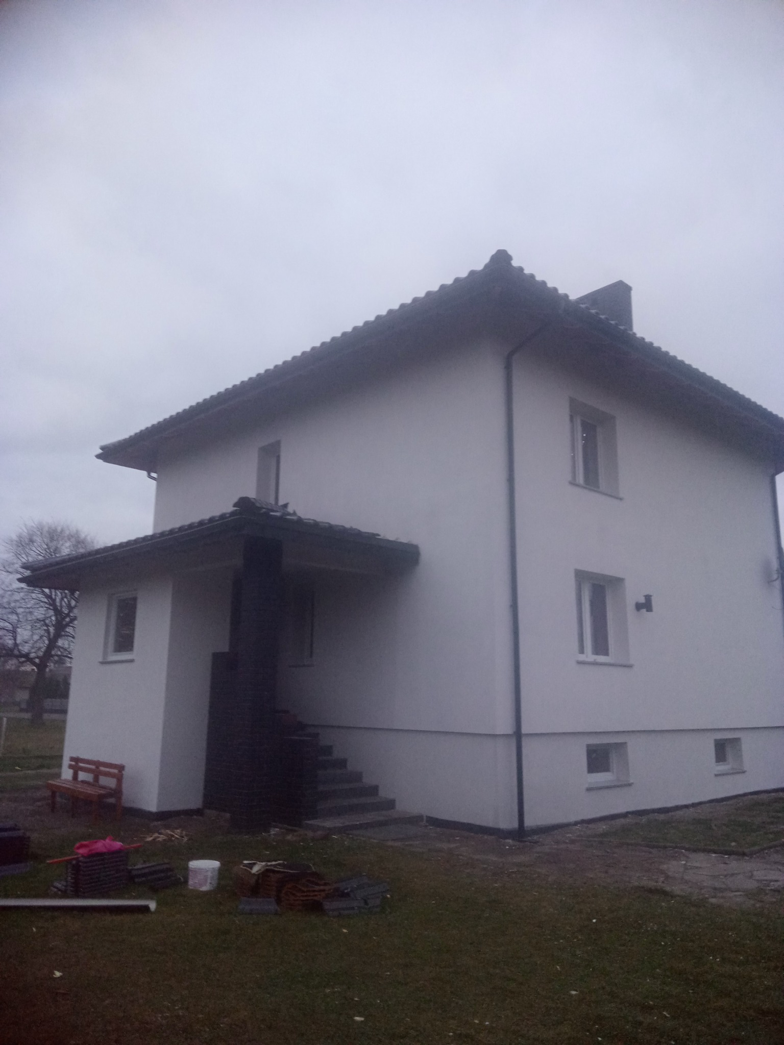 Dwukondygnacyjny dom z jasną elewacją po renowacji, widoczne pozostałości materiałów budowlanych i drewniana ławka przed wejściem.