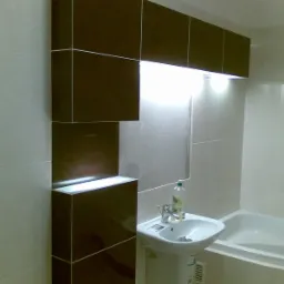 Wykończona łazienka z brązowymi i białymi płytkami, podświetlanymi półkami i umywalką na postumencie, widoczna butelka płynu do mycia.