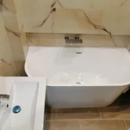 Wykończona łazienka z beżowymi płytkami imitującymi marmur, białą wanną wolnostojącą i umywalką z chromowaną baterią.