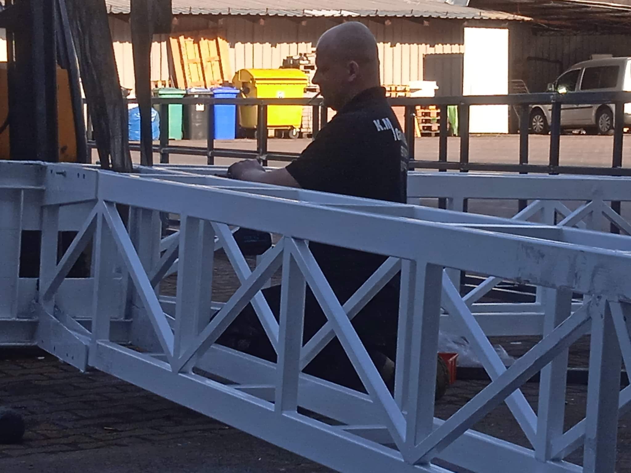 Pracownik w czarnej koszulce z logo firmy siedzi na pomalowanej na biało konstrukcji kratownicowej, prawdopodobnie w trakcie przerwy. W tle widoczne pojemniki na śmieci i budynek magazynowy.