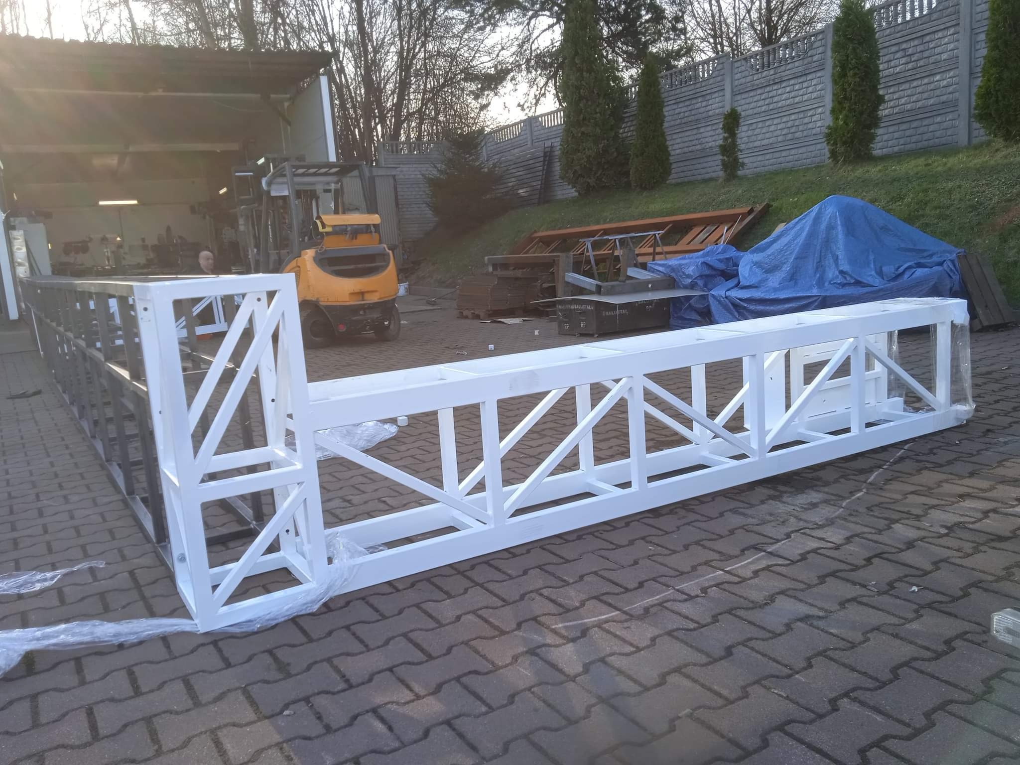 Fragment konstrukcji kratownicowej pomalowanej na biało, wykonanej ze stali, częściowo owiniętej folią, leżącej na brukowanej powierzchni, z wózkiem widłowym w tle.
