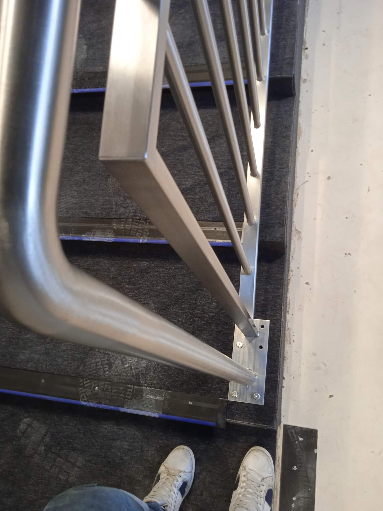 Szczegółowe ujęcie z góry na zamontowaną balustradę nierdzewną na schodach z szarego dywanu, widoczne mocowania śrubowe i fragment butów osoby fotografującej w dolnej części kadru.