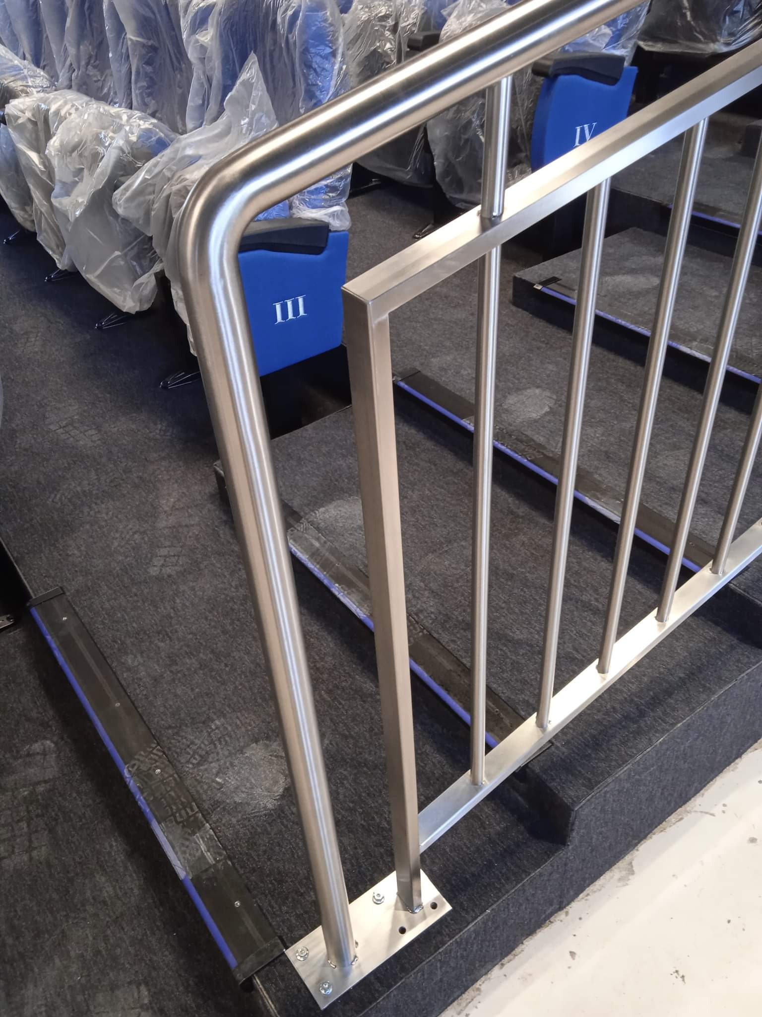 Metalowa barierka schodowa z rur, zabezpieczająca rzędy siedzeń w kinie lub teatrze, z widocznymi numerami rzędów na siedzeniach.