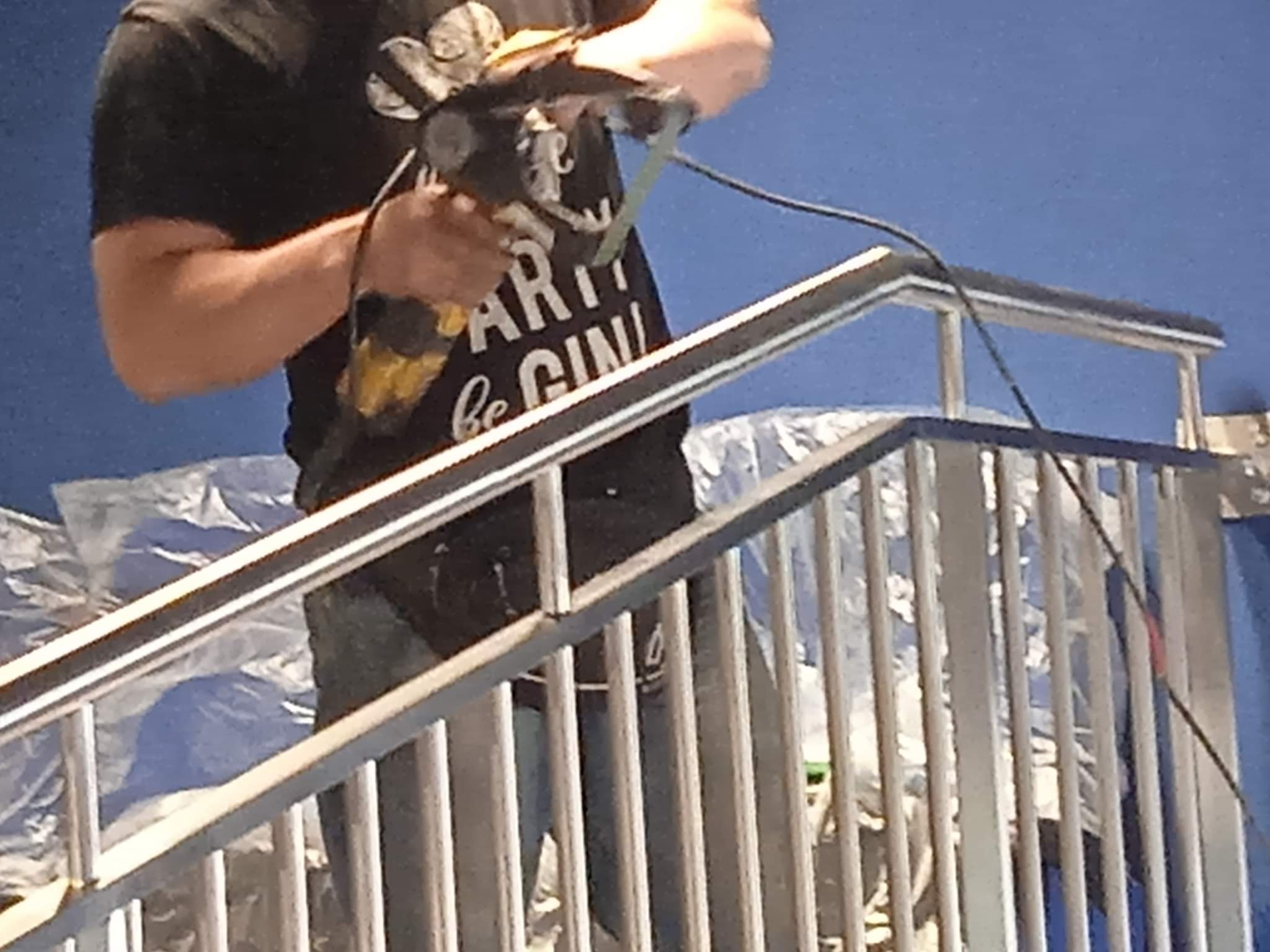 Pracownik w czarnej koszulce z nadrukiem szlifuje spaw na metalowej konstrukcji schodów, widoczne zabezpieczenia folią malarską na niebieskim tle.