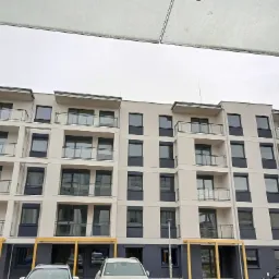 Wielopiętrowy budynek mieszkalny z balkonami, świeżo pomalowany na biało i grafitowo, widoczny z dołu, z fragmentem dachu samochodu na pierwszym planie i pochmurnym niebem w tle.