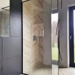 Nowoczesna kabina prysznicowa walk-in z czarną ramą, wykończona płytkami w jodełkę, z widoczną armaturą i odpływem liniowym.