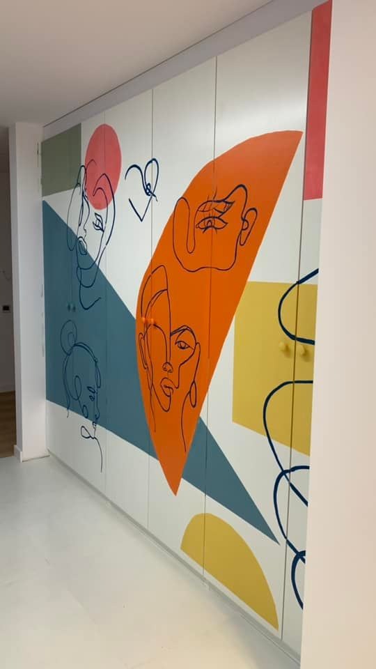 Wielobarwna szafa wnękowa z abstrakcyjnymi, linearnymi portretami i geometrycznymi kształtami w odcieniach pomarańczy, błękitu, zieleni i żółci, na białym tle, z okrągłymi uchwytami.