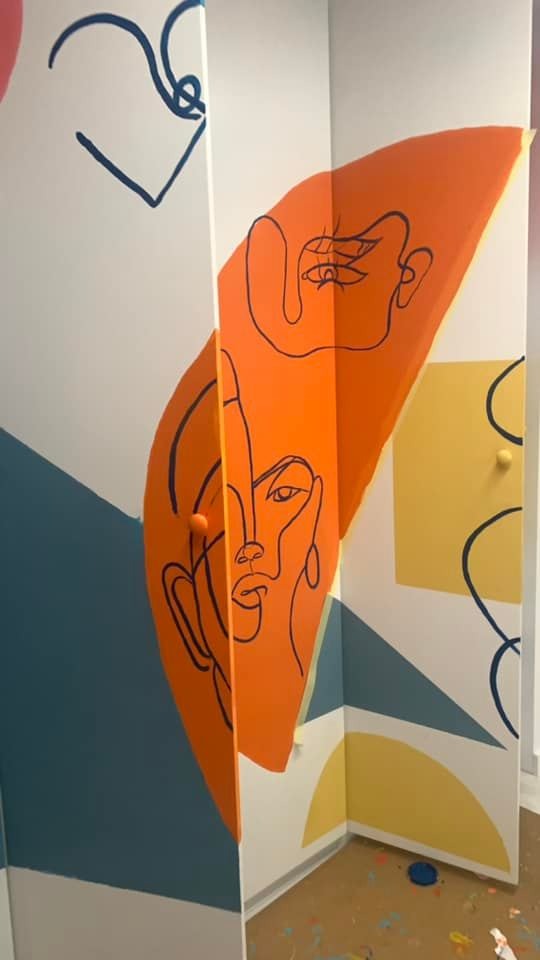 Abstrakcyjne malowidło ścienne w pokoju z motywami twarzy narysowanych czarną linią na pomarańczowym tle, uzupełnione geometrycznymi kształtami w odcieniach niebieskiego i żółtego.