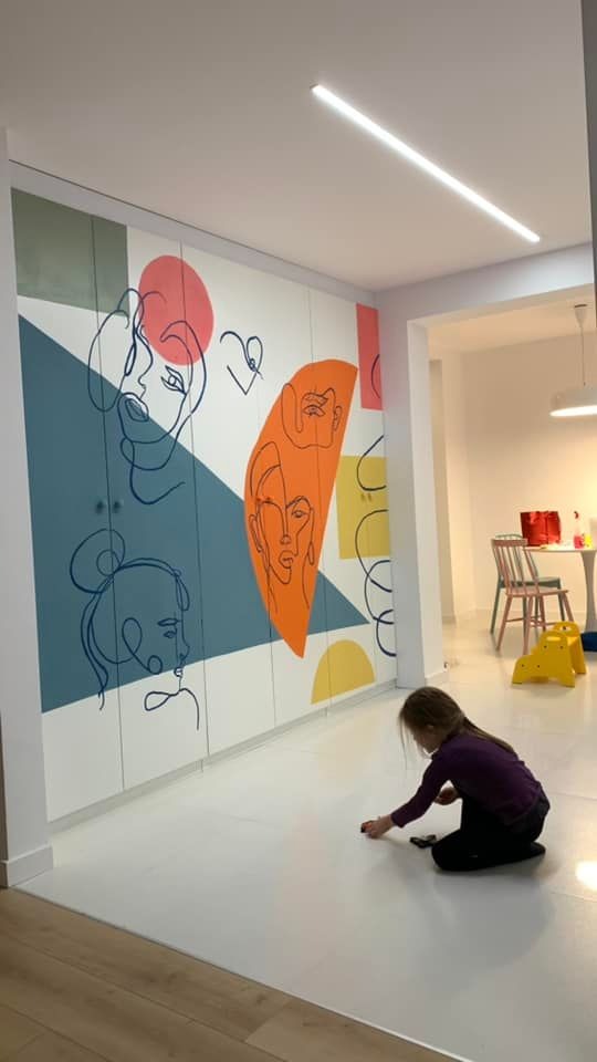 Wnętrze pokoju z szafą w zabudowie, ozdobioną abstrakcyjnymi rysunkami twarzy w kolorach niebieskim, pomarańczowym i różowym. Dziecko bawi się na jasnej podłodze, w tle widoczny stół i krzesła.