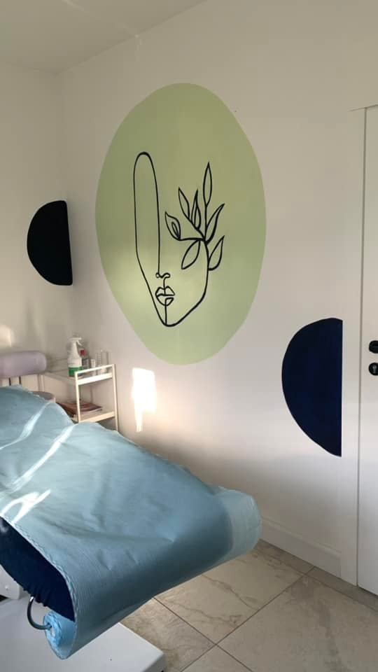 Wnętrze gabinetu kosmetycznego z łóżkiem zabiegowym; na ścianie minimalistyczny rysunek twarzy i liści w zielonym okręgu oraz dwa półkola w kolorze granatowym.