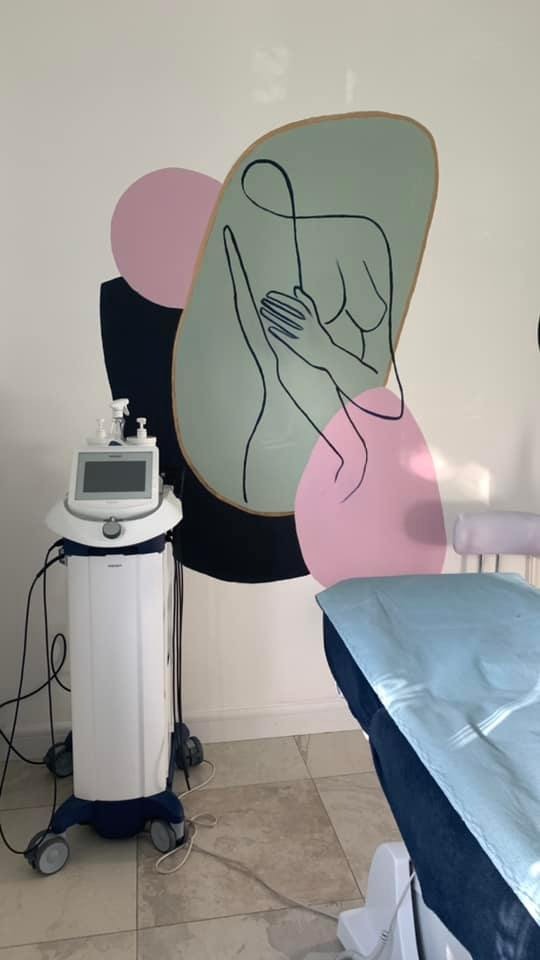 Wnętrze gabinetu kosmetycznego z aparaturą do zabiegów i minimalistycznym malowidłem ściennym przedstawiającym kobiecą sylwetkę w odcieniach pastelowych różów, zieleni i czerni.