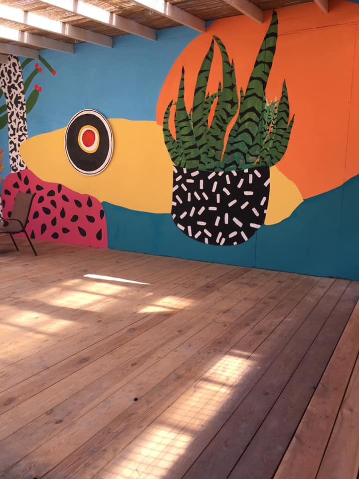 Barwna, abstrakcyjna kompozycja malarska na ścianie zewnętrznej budynku, przedstawiająca stylizowaną roślinę w doniczce na tle geometrycznego słońca, z drewnianym tarasem w ciepłych odcieniach...