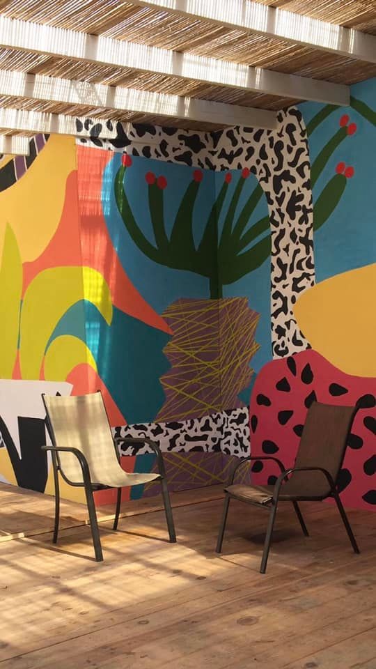 Dwa krzesła na drewnianej podłodze przed ścianą pokrytą abstrakcyjnym, kolorowym malunkiem z motywami roślinnymi i geometrycznymi, pod ażurowym zadaszeniem.