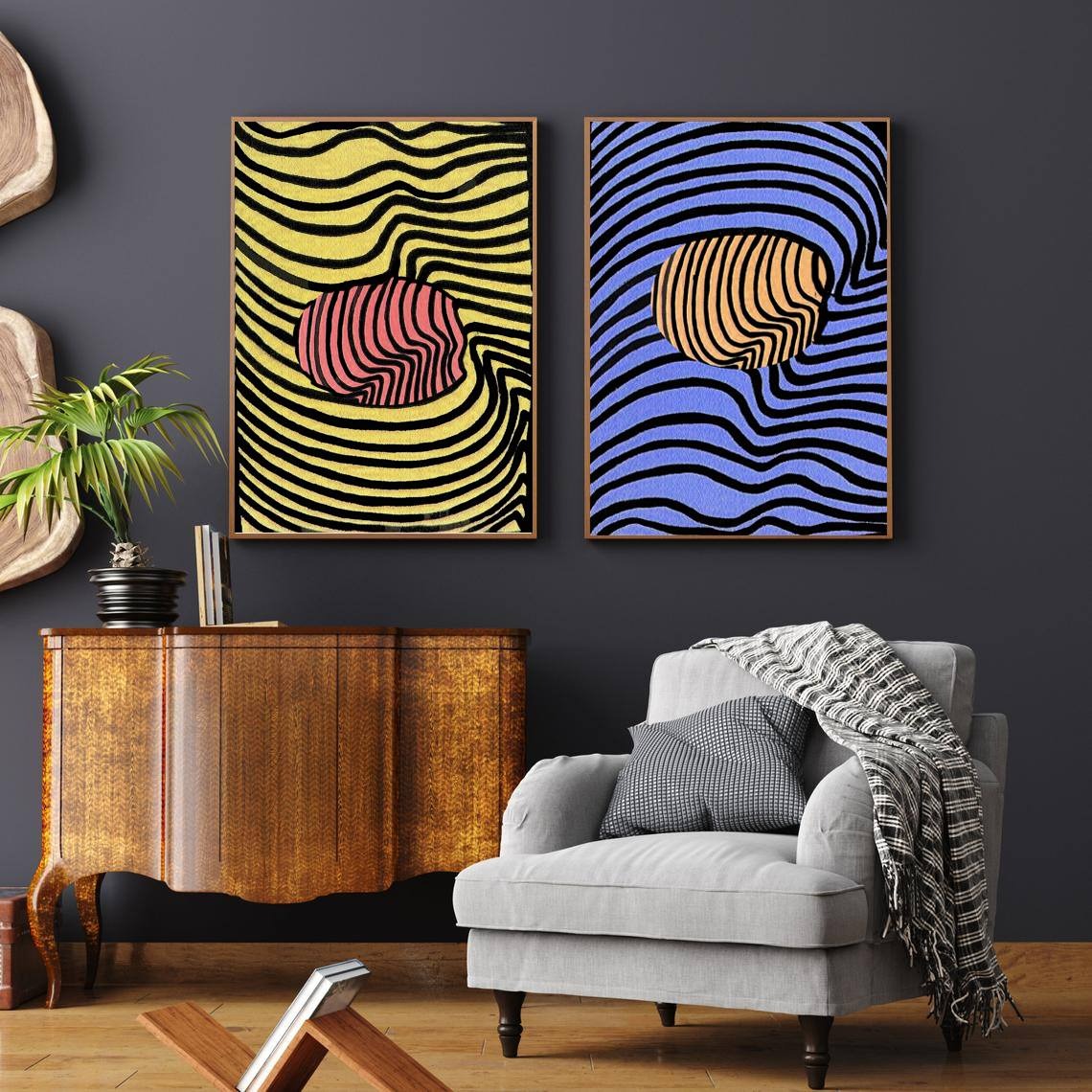 Dwa abstrakcyjne obrazy w ramach wiszące nad komodą i fotelem. Jeden obraz z żółtym tłem i różowym kształtem, drugi z niebieskim tłem i pomarańczowym kształtem. Obrazy w nowoczesnym salonie...