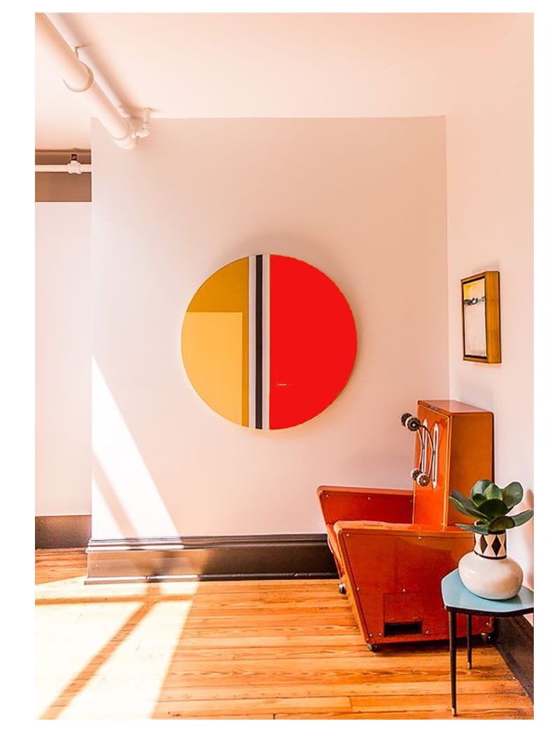 Wnętrze z okrągłym obrazem w kolorach żółci i czerwieni na ścianie, obok stoi pomarańczowy fotel vintage i niebieski stolik z rośliną doniczkową.