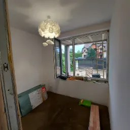 Pokój w trakcie remontu: widoczne przygotowane panele podłogowe VOX MaxPod, listwy przypodłogowe, lampa sufitowa w kształcie kwiatu oraz okno z widokiem na dom jednorodzinny.