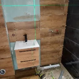 Niedokończona łazienka z drewnopodobnymi płytkami ściennymi i podłogowymi, ustawiona szafka umywalkowa z czarną armaturą, poziomica laserowa wyznaczająca linie, widoczne narzędzia i akcesoria...