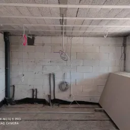 Surowy stan wnętrza w trakcie prac: ściany z białego bloczka, betonowy sufit z wystającymi przewodami elektrycznymi, rury kanalizacyjne, narzędzia przy ścianie oraz płyty gipsowo-kartonowe przy oknie.