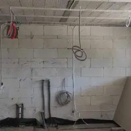 Surowy stan wnętrza z białych bloczków, widoczne przewody elektryczne zwisające z sufitu, rury i płyty kartonowo-gipsowe, okno w tle.