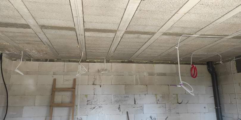 Surowy betonowy strop z widocznymi belkami i instalacjami elektrycznymi w trakcie montażu, ściany z białych bloczków, drewniana drabina oparta o ścianę.