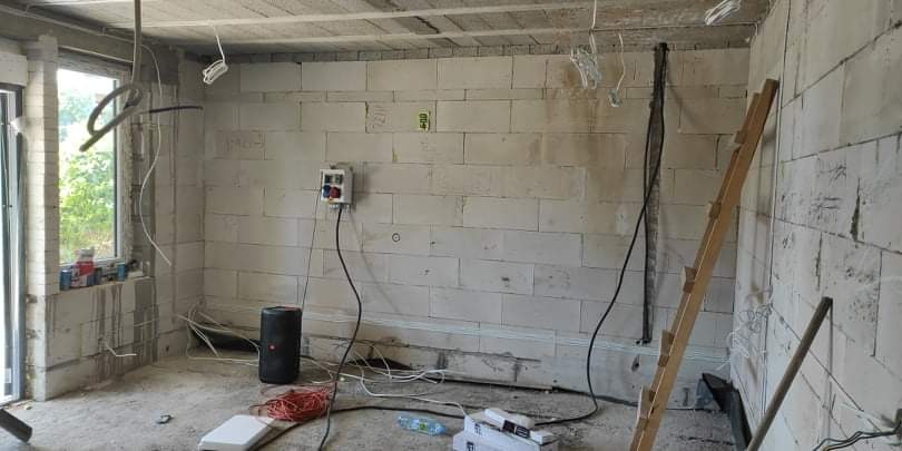 Surowy stan pomieszczenia z bloczków, widoczne przewody elektryczne, puszka z bezpiecznikami na ścianie, drabina oparta o ścianę, okno z widokiem na zieleń za nim.