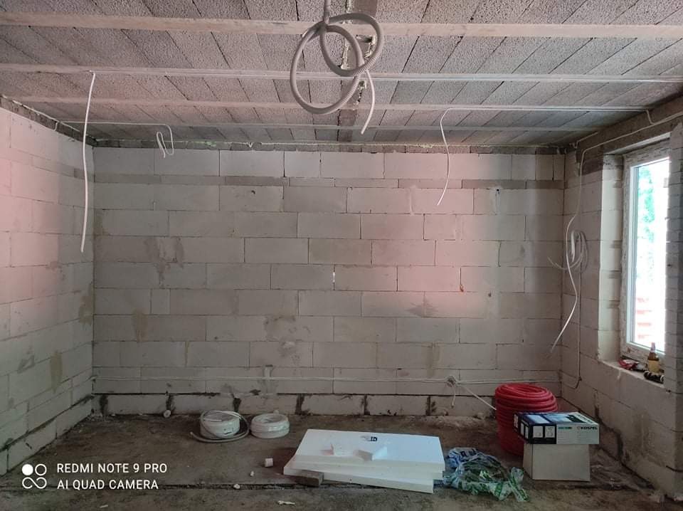 Surowy pokój z białego bloczka, widoczne przewody elektryczne wystające ze ścian i sufitu, przygotowane materiały izolacyjne na podłodze, okno z widokiem na zieleń.