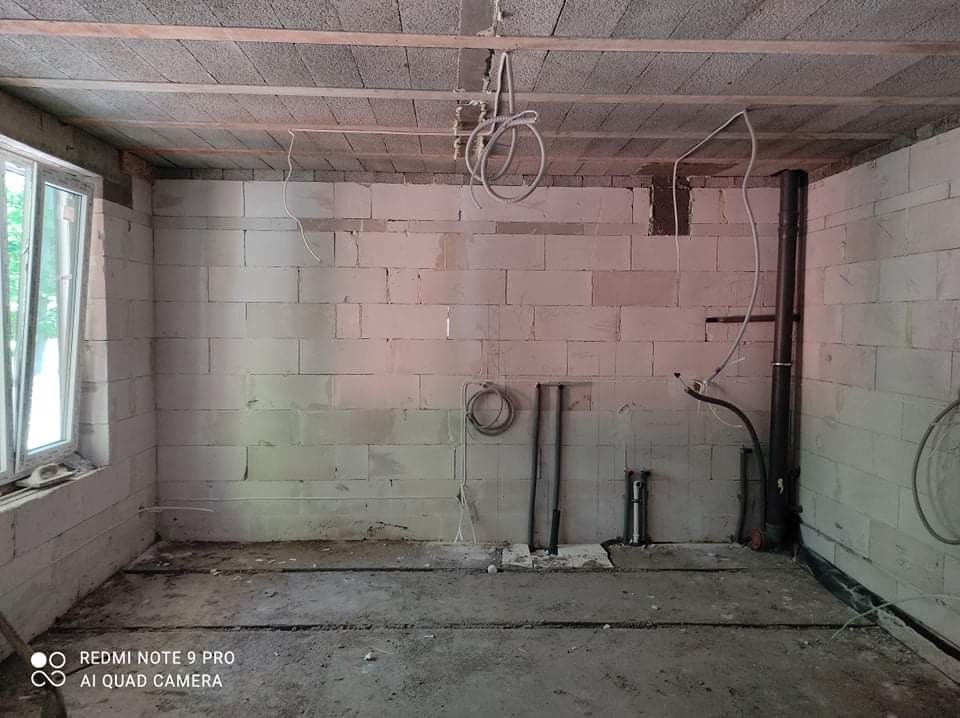 Surowy stan pomieszczenia w trakcie prac: ściany z białych bloczków, widoczne przewody elektryczne zwisające z sufitu, rury pionowe przy ścianie, częściowo przygotowana podłoga, okno z widokiem...