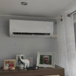 Biały klimatyzator ścienny w pokoju z szarymi ścianami, wiszący nad komodą z ramkami ze zdjęciami i dekoracyjną figurką słonia, widoczny fragment telewizora i okno z roletami.