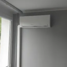 Nowoczesny, biały klimatyzator ścienny zamontowany na szarej ścianie w pokoju z widokiem na zieleń za oknem.