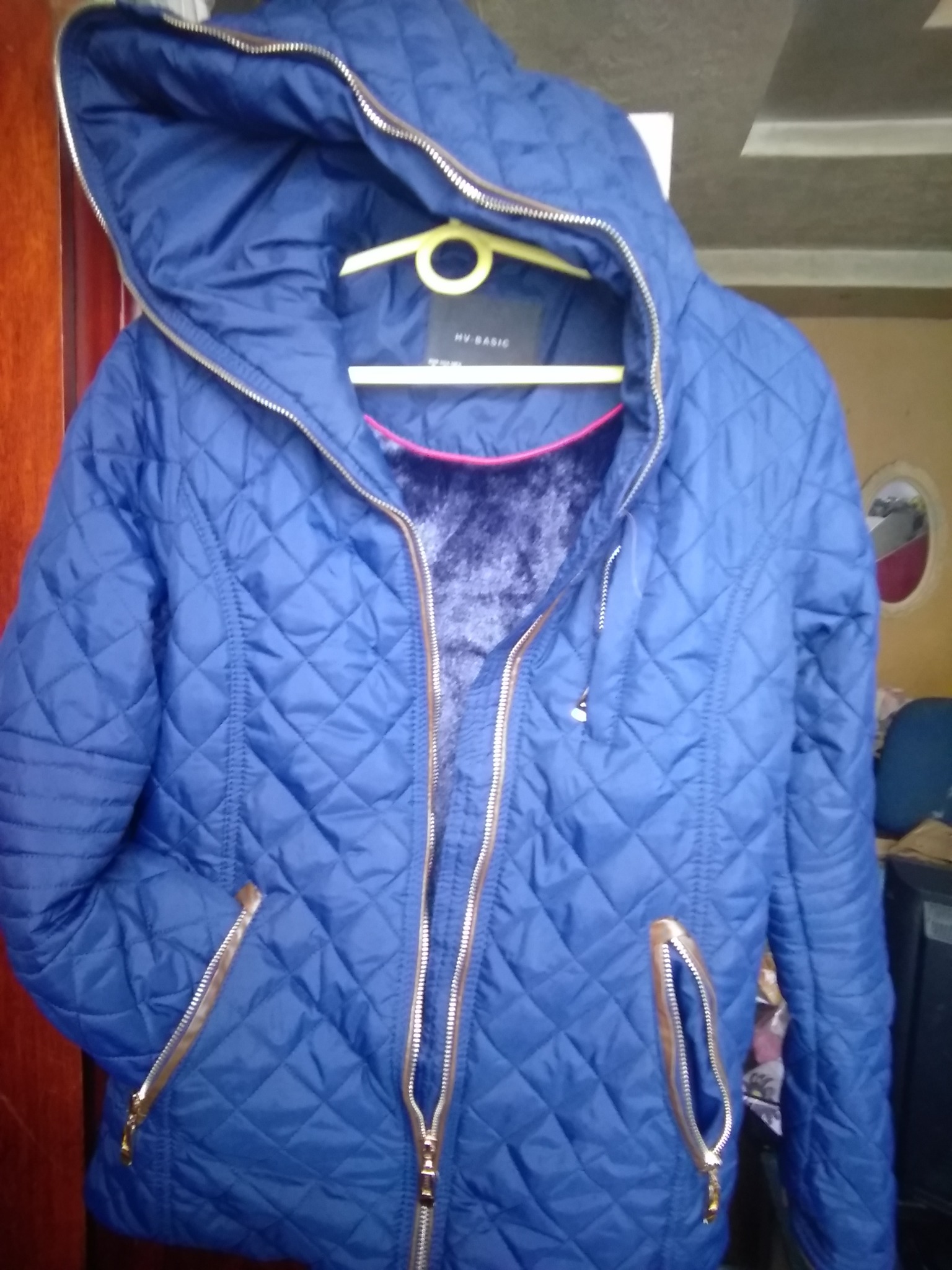 Niebieska, pikowana kurtka damska z kapturem, wisząca na żółtym wieszaku, z widoczną podszewką z futerka i złotymi zamkami błyskawicznymi.