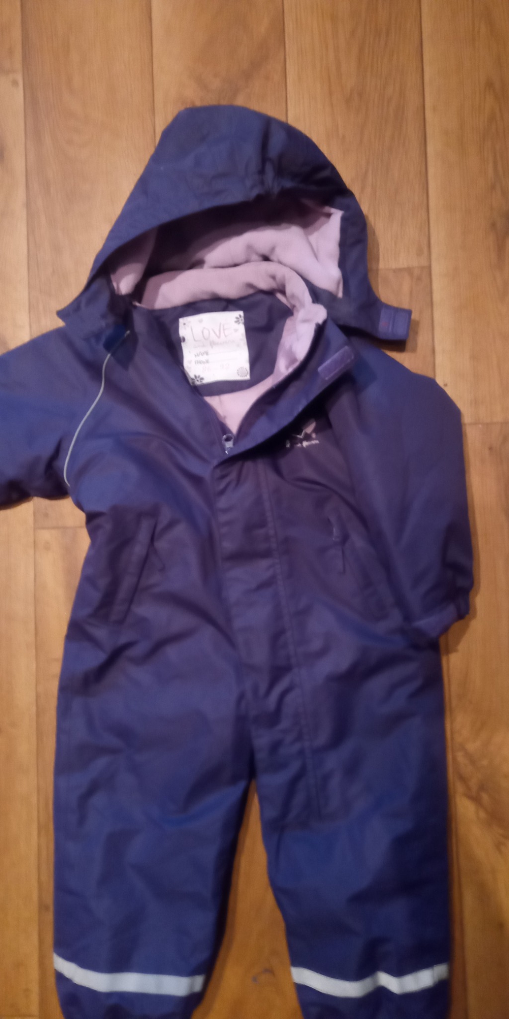 Niebieski kombinezon dziecięcy z kapturem, rozłożony na drewnianej podłodze, widoczna metka z napisem 'LOVE' i logo marki.