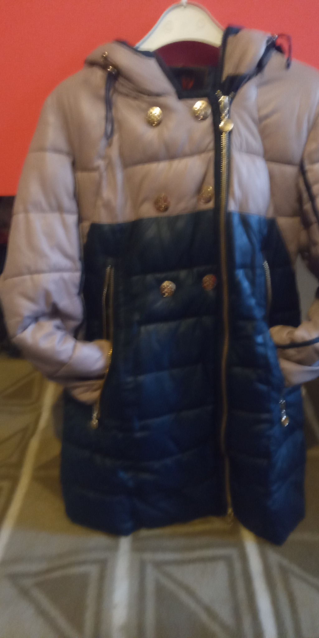 Dwukolorowa, pikowana kurtka damska z kapturem, zapinana na zamek i zatrzaski z ozdobnymi złotymi guzikami, wisząca na wieszaku.