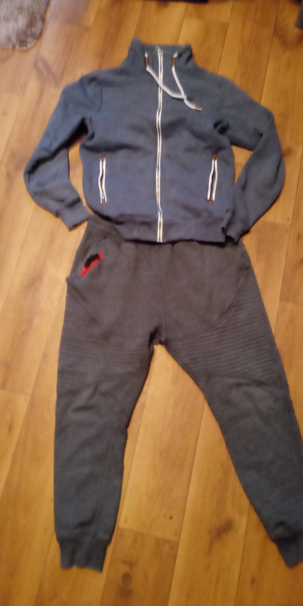 Szary dres męski z bluzą na zamek i spodniami dresowymi z poziomymi przeszyciami na kolanach, leżący na drewnianej podłodze.