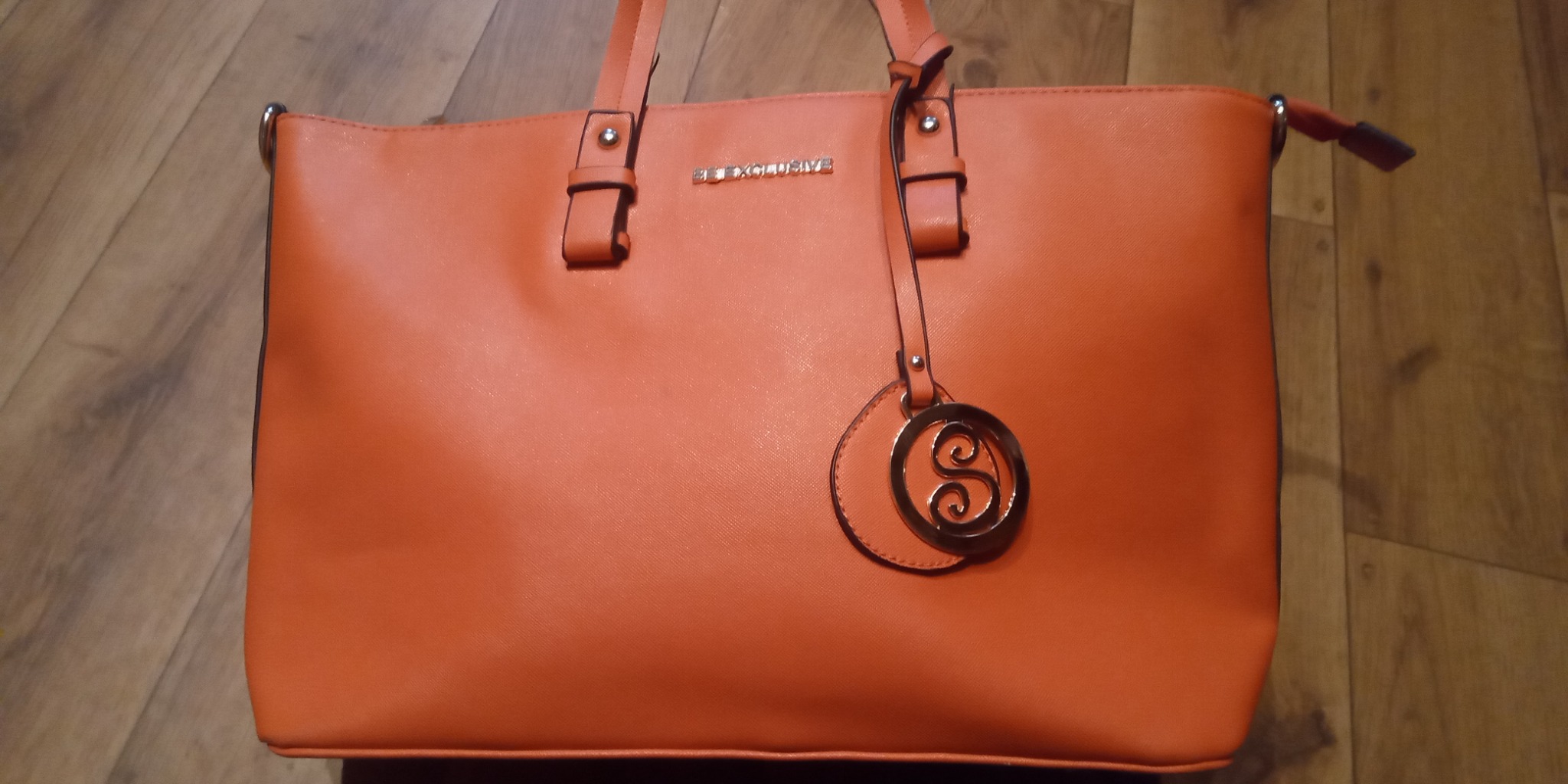 Pomarańczowa torebka damska z logo 'BE EXCLUSIVE' i okrągłym ozdobnym brelokiem na tle drewnianej podłogi.