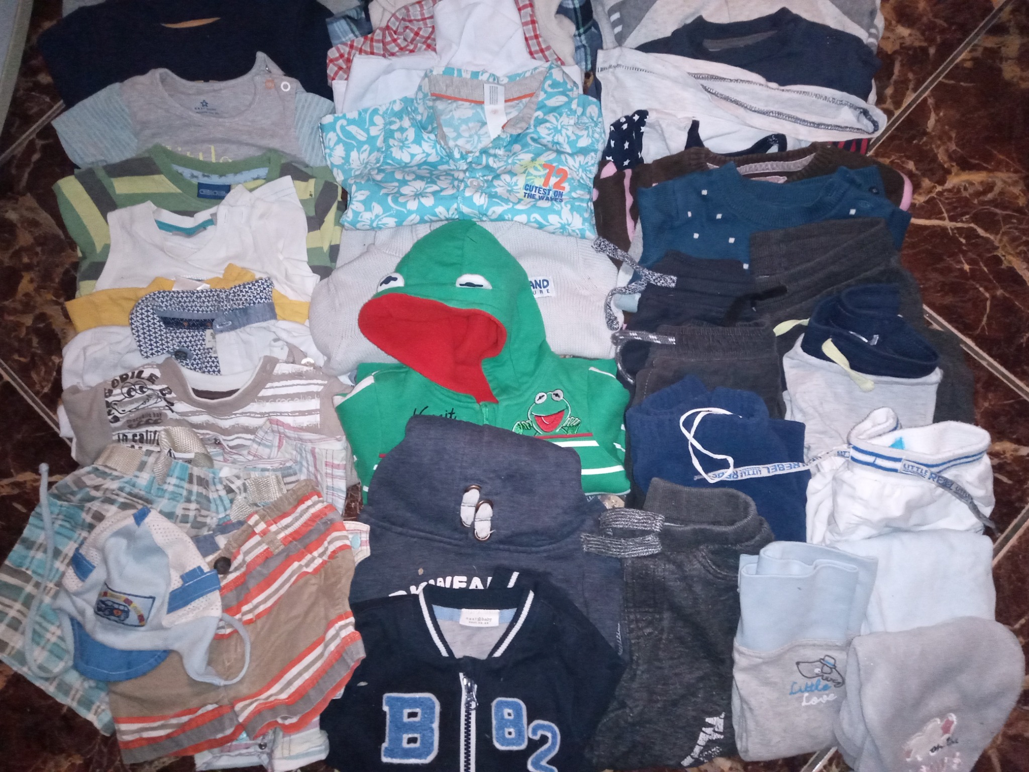 Stos różnorodnych ubrań dziecięcych, w tym bluzki z krótkim rękawem, bluzy z kapturem (jedna z motywem żaby), koszule i spodenki, ułożonych na brązowej podłodze z wzorem marmuru.