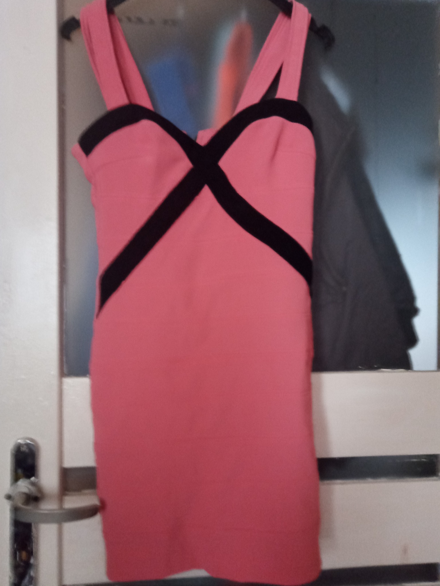 Różowa sukienka bandażowa z czarnymi akcentami, wisząca na wieszaku przed drzwiami.