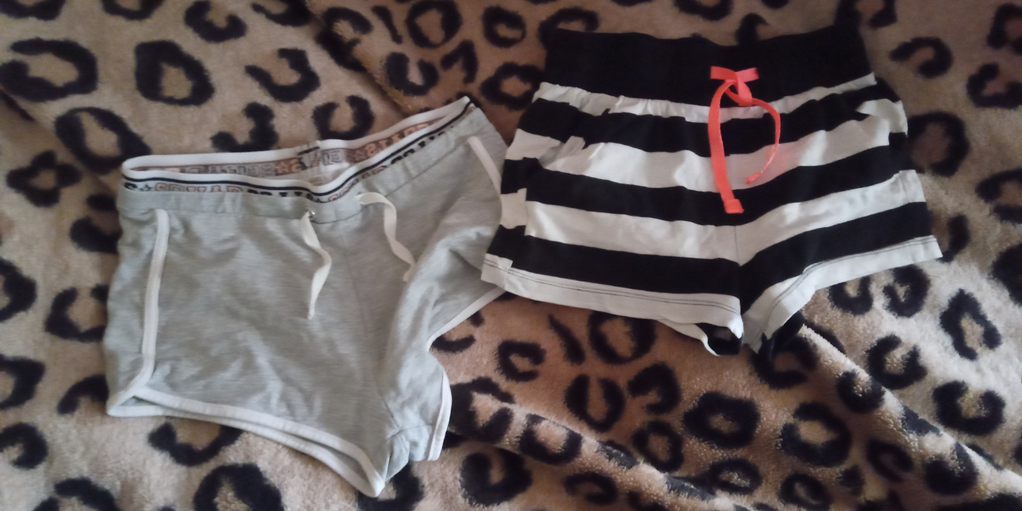 Dwie pary krótkich spodenek damskich: szare sportowe z białym wykończeniem i czarno-białe w paski z pomarańczową kokardką, leżące na kocu w panterkę.