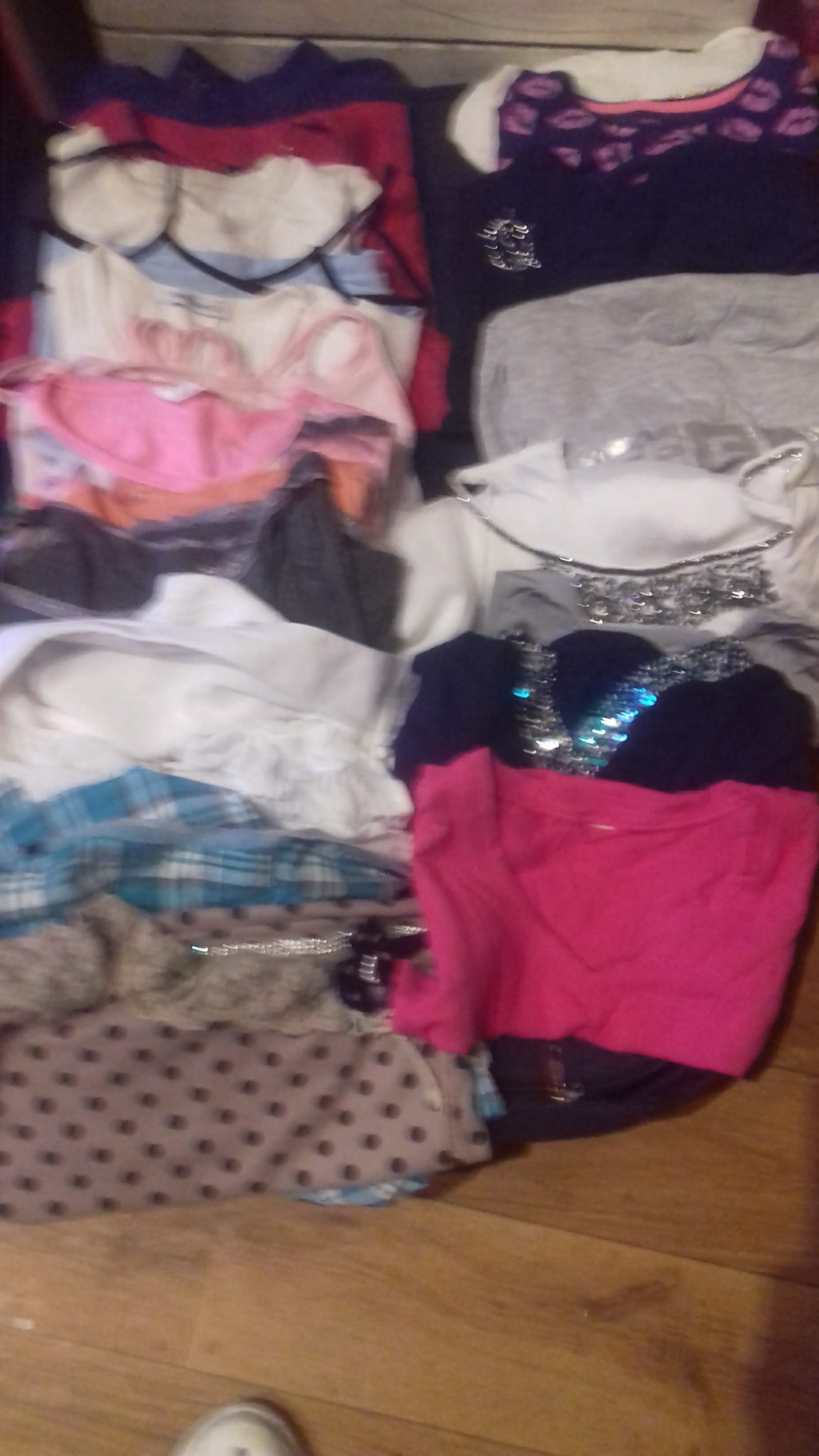 Stos ubrań damskich z różnymi wzorami i teksturami, w tym bluzki z cekinami, koszulki w kropki i kraciaste koszule, ułożonych na drewnianej podłodze.