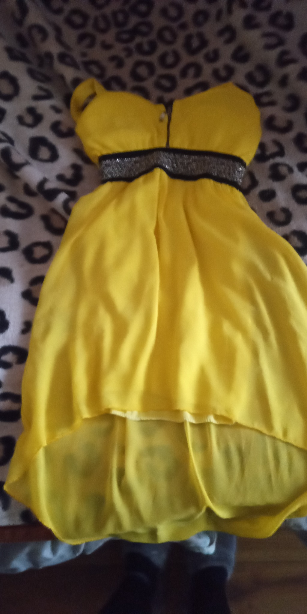 Żółta, zwiewna sukienka z asymetrycznym dołem i srebrnym paskiem, leżąca na wzorzystym tle.