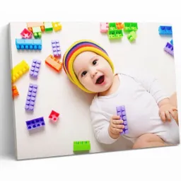 Uśmiechnięte niemowlę w kolorowej czapce leży na białym tle otoczone kolorowymi klockami, trzymając jeden w dłoni, wydruk na płótnie.