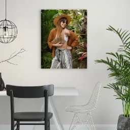 Portret uśmiechniętej kobiety w kapeluszu i okularach przeciwsłonecznych, pozującej z torebką na tle tropikalnej roślinności, widoczny jako obraz na ścianie w nowoczesnym wnętrzu z krzesłami...