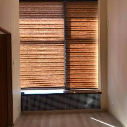 Wnętrze pokoju z drewnianą podłogą i brązowymi żaluzjami w oknie, osłaniającymi grzejnik z metalową kratką, ściany w odcieniach beżu i kremu, widoczny fragment brązowych drzwi po lewej stronie.