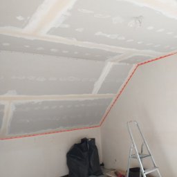 Kamil Siwek - Skośne poddasze w trakcie montażu płyt gipsowo-kartonowych, widoczne profile, szpachlowane łączenia, drabina, narzędzia i worki na odpady budowlane.