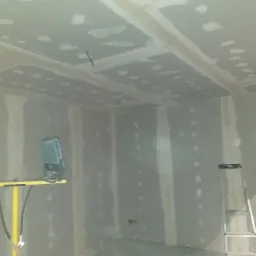 Montaż płyt kartonowo-gipsowych na suficie i ścianach, widoczne łączenia wzmocnione taśmą, oświetlenie halogenowe na żółtym statywie i aluminiowa drabina w pomieszczeniu w trakcie remontu.