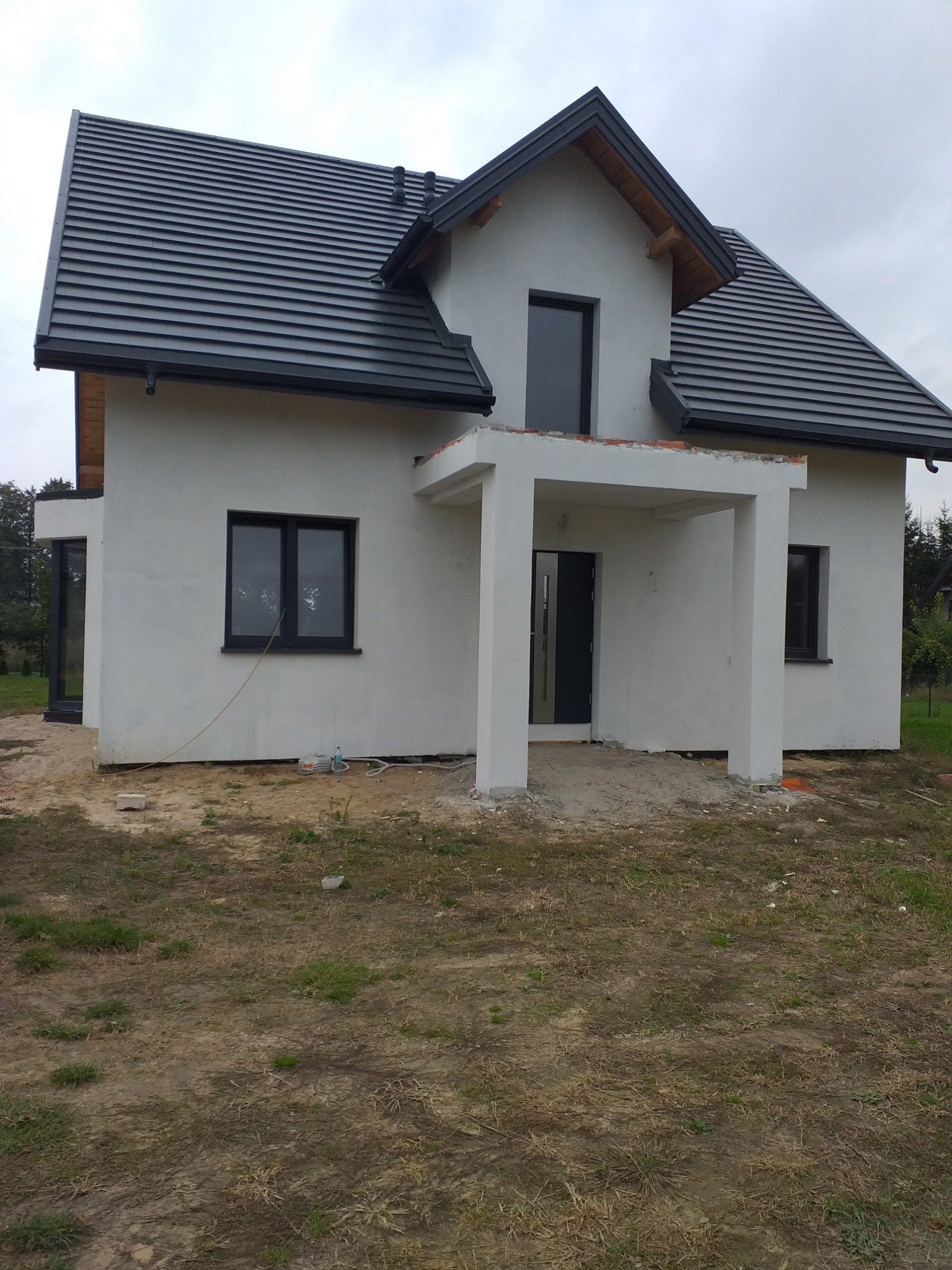 Nowo wybudowany dom jednorodzinny z szarą dachówką i białymi ścianami, widoczne niedokończone prace wykończeniowe w otoczeniu.
