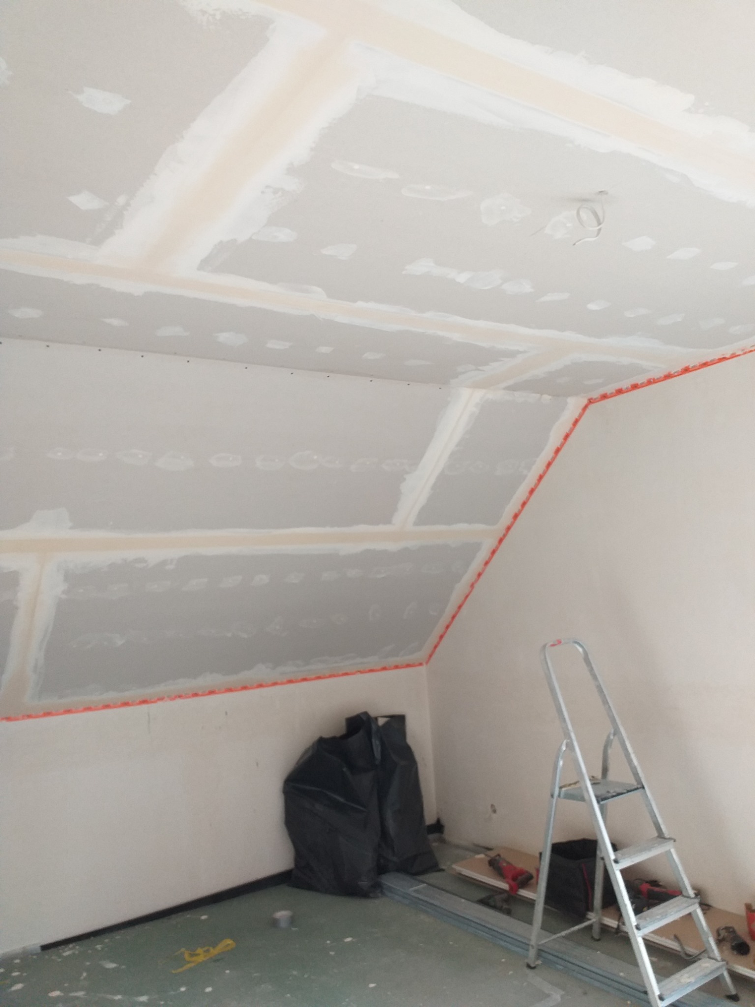 Skośne poddasze w trakcie montażu płyt gipsowo-kartonowych, widoczne profile, szpachlowane łączenia, drabina, narzędzia i worki na odpady budowlane.