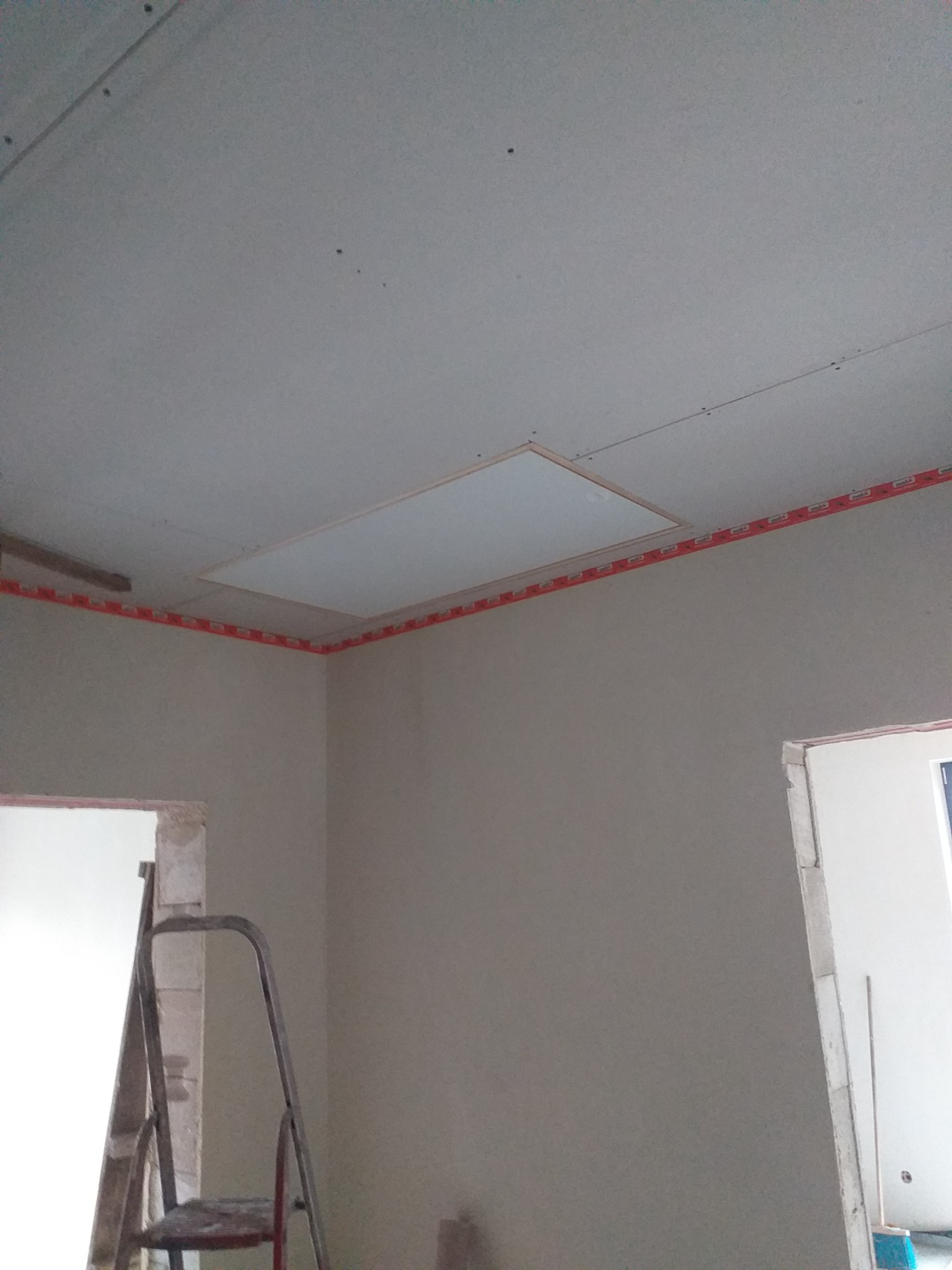 Wnętrze w trakcie prac wykończeniowych: sufit z zamontowaną płytą rewizyjną, ściany przygotowane do malowania, widoczna drabina i zabezpieczenia narożników.