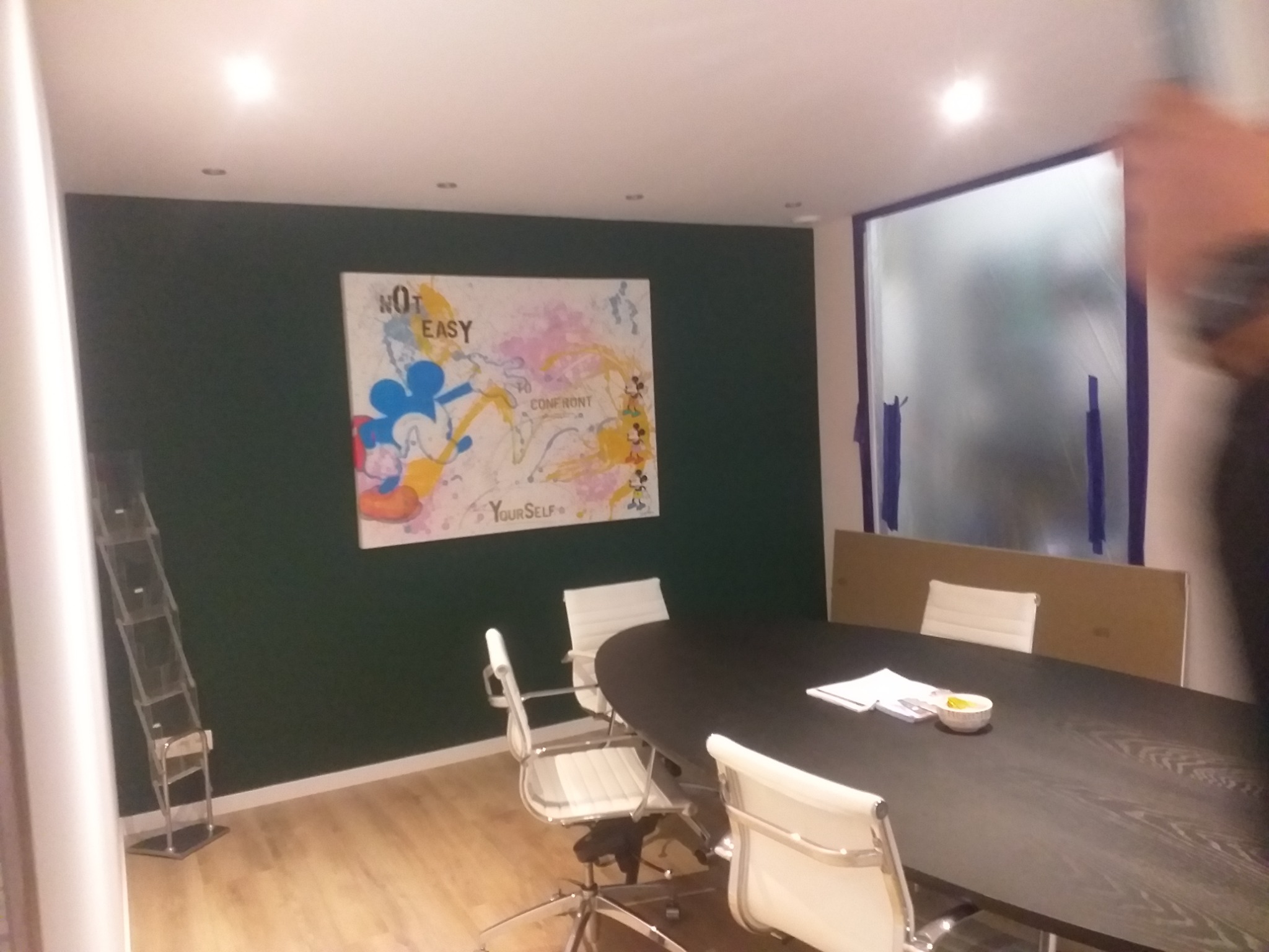 Biuro po malowaniu ścian na ciemnozielony kolor, z zabezpieczonym oknem folią malarską, abstrakcyjnym obrazem na ścianie i czarnym stołem konferencyjnym.