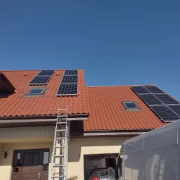 Instalacja paneli słonecznych na dachu domu z czerwoną dachówką, widoczna drabina i częściowo samochód dostawczy.